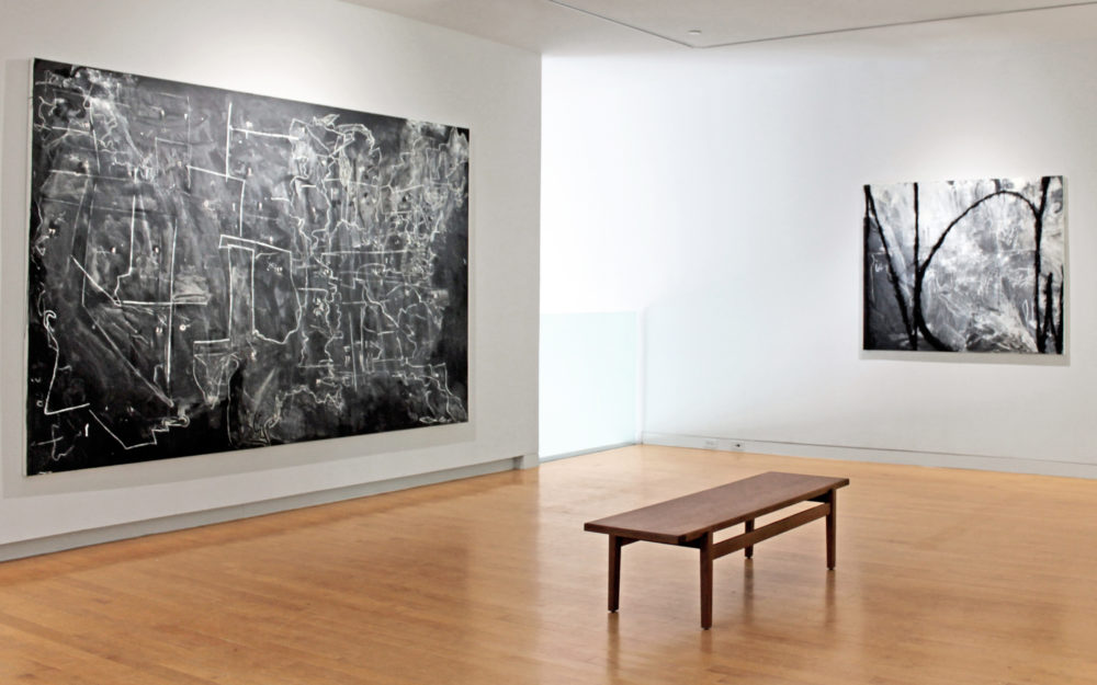 Install view of a gallery with a painting of an abstract map of the United States to the left, a bench in the center and a smaller abstract work to the right.