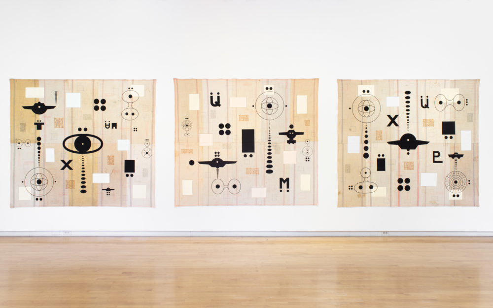 Three large-scale unstretched paintings with similar dark motifs against cream colored background hung together on a wall.