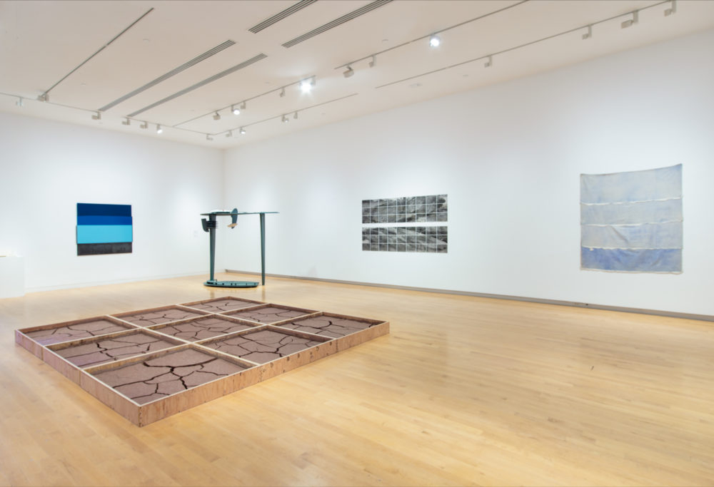 Gallery with paintings on the wall and two abstract sculptures, one tall and made of dark metal, the other on the floor, a wooden grid filled with red clay.