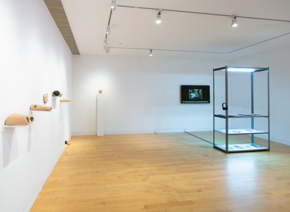 Small sculptural pieces on the wall to the left in the shape of body parts and an illuminated shelf with objects and video projection at right.
