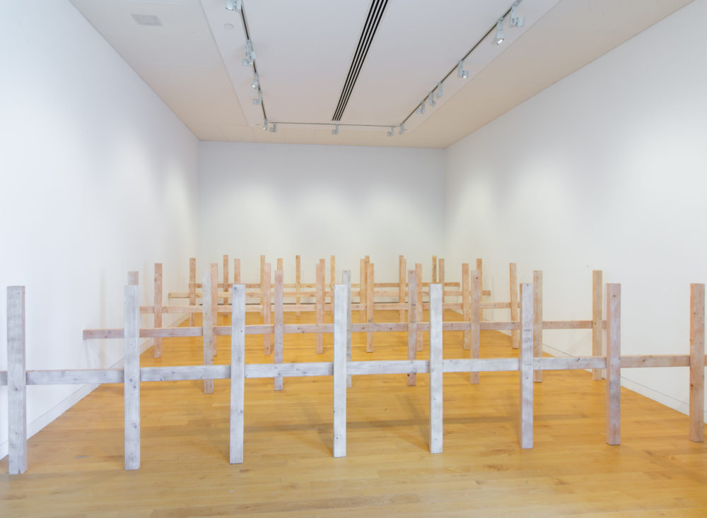 Several unpainted wooden fences in a row that fill a gallery space.
