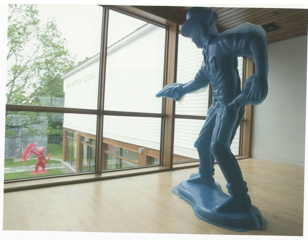 Large bright blue plastic Cowboy figure in gallery aiming gun at red Indian installed outside on Museum lawn.