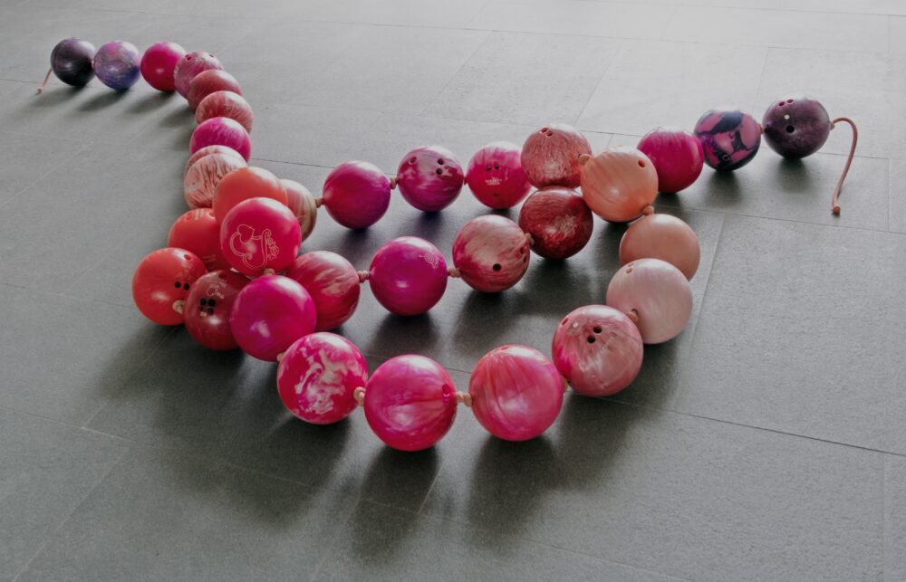 A large scale sculpture of a string of pearls made of bright pink bowling balls coiled on a dark tile floor