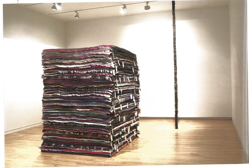 Right to Left: Tall pile of wool blankets; thin bronze casted items stacked into a column in gallery..