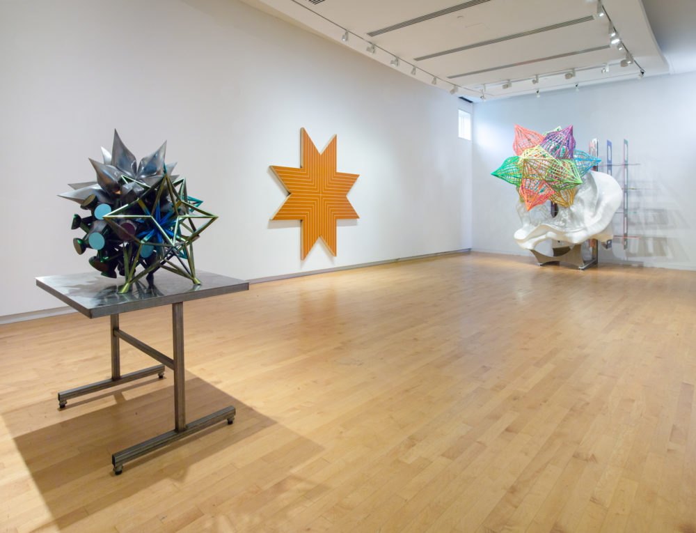 Photo of gallery with a small sculpture to left, a large star-shaped painting at center, and a large sculpture featuring a colorful star to the right.
