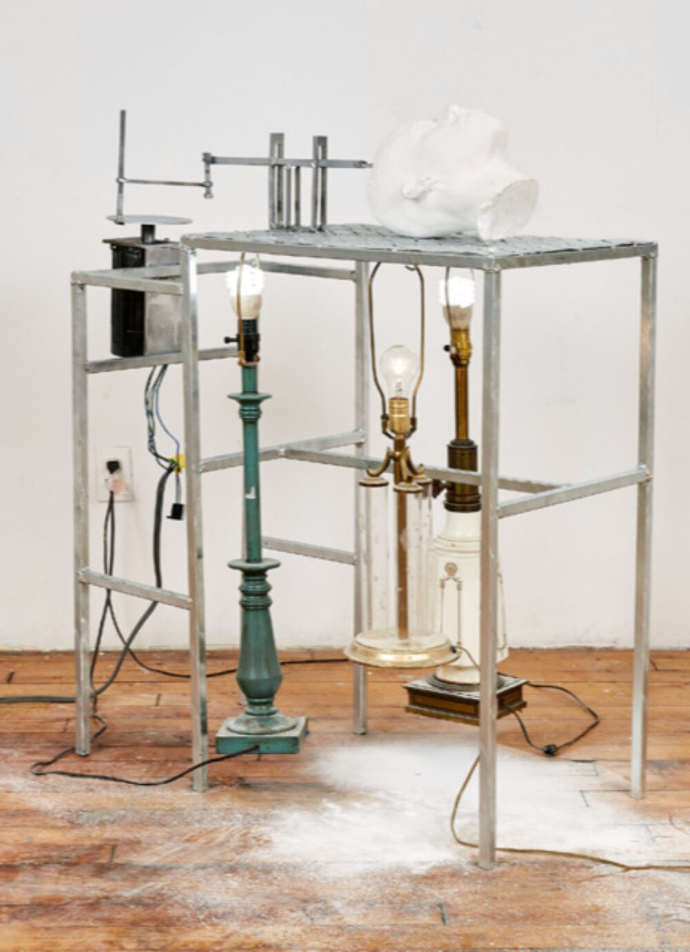 Metal table base with lamps and a white cast human head that's being grated into dust collecting on the floor.