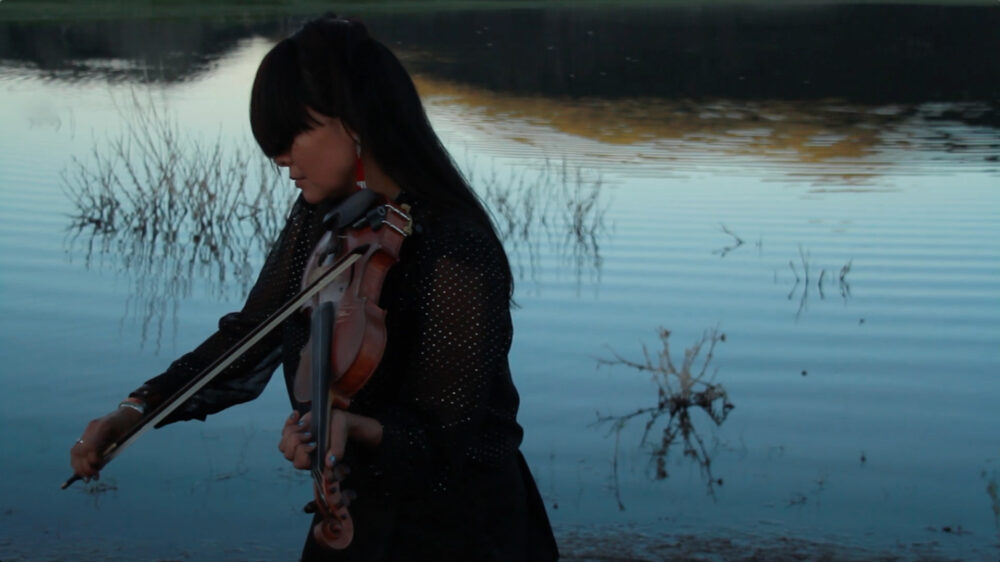 Artist Laura Ortman playing the violin in front of a body of water at dusk