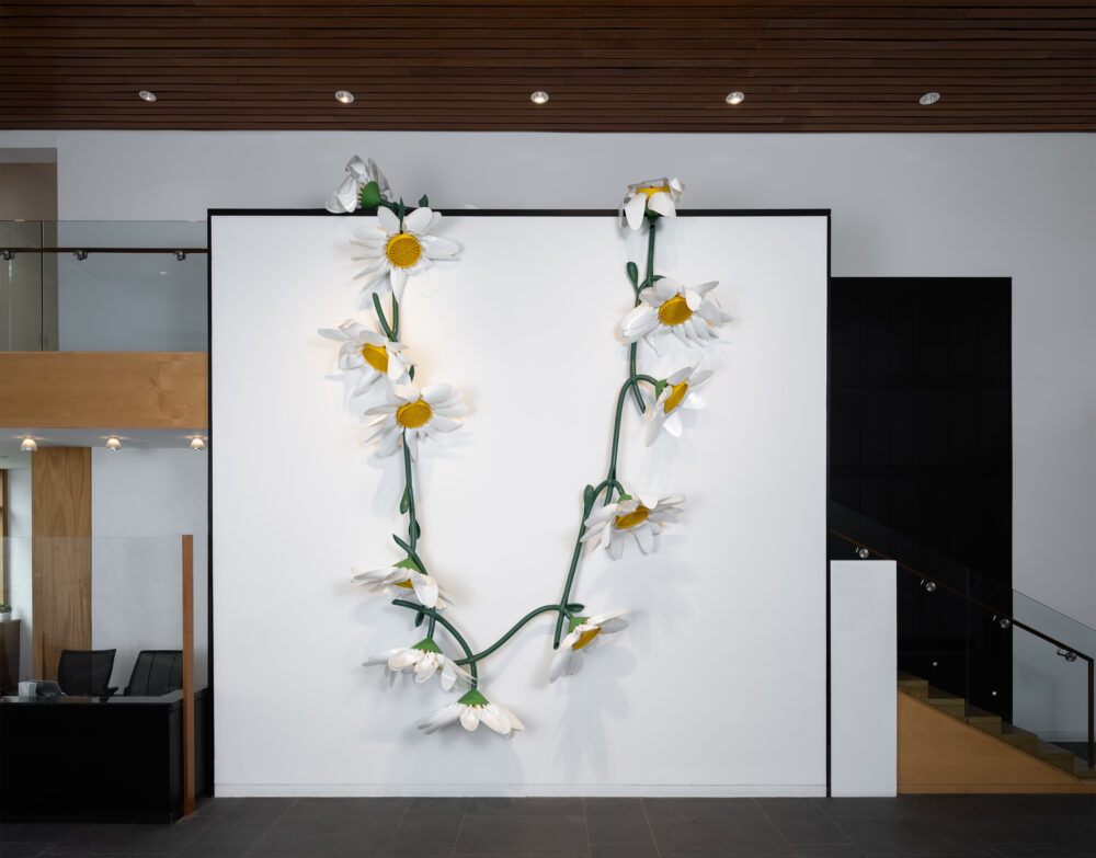 A large scale sculpture of a daisy chain made of plastic, and gardening materials hanging over a white wall in a museum