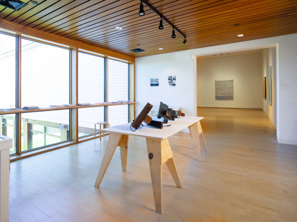 Gallery on an indoor bridge with large windows and a table with artworks in the center along with tables with a library of archival material and books