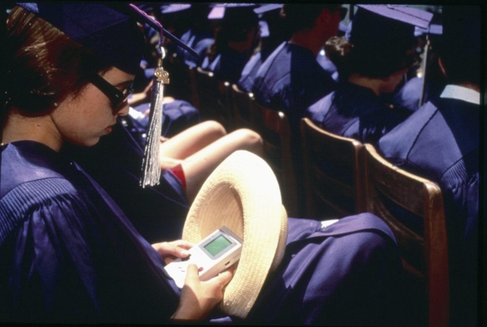 A girl sitting at graduation, with her cap and gown, playing on a Gameboy in secret
