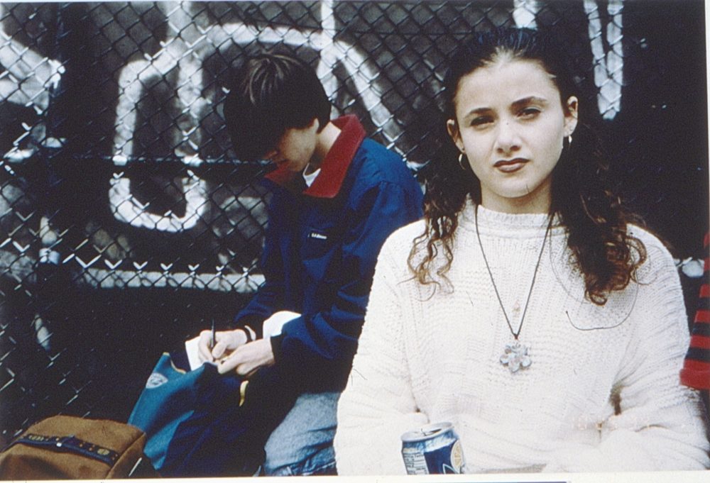 This is a photograph of two young teens. The young boy is wearing a blue windbreaker and the girl looking at the camera is in a white sweater