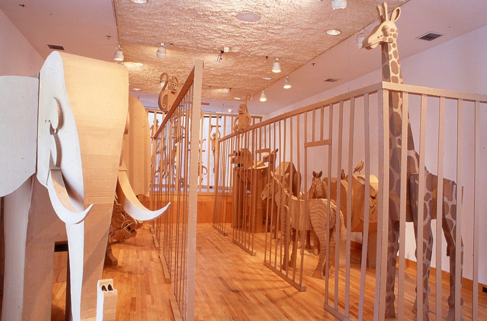 A cardboard elephant and giraffe amongst other animals behind a cardboard cage