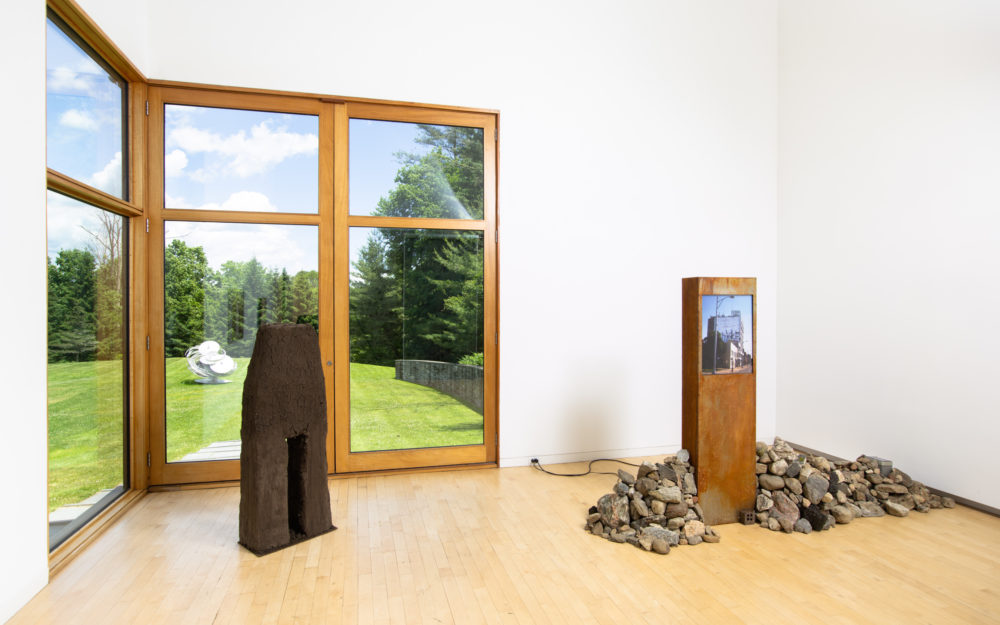 Two freestanding sculptural works, one with a pile of stones, the other made of earth. Through the window there's a silver sculpture outside.