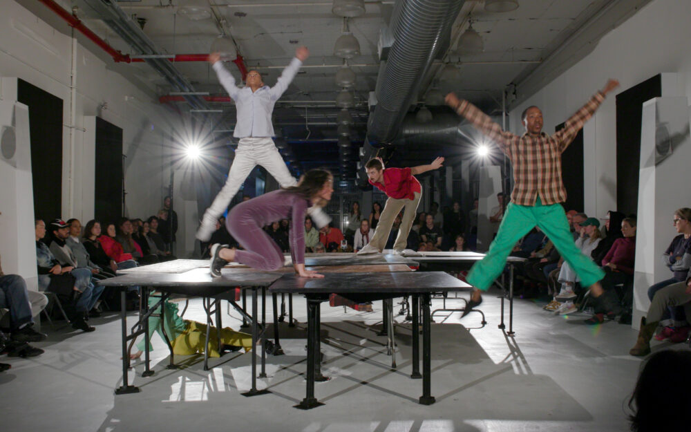 Performers standing, crouching and jumping off and on tables in a room with an audience around the perimeter.