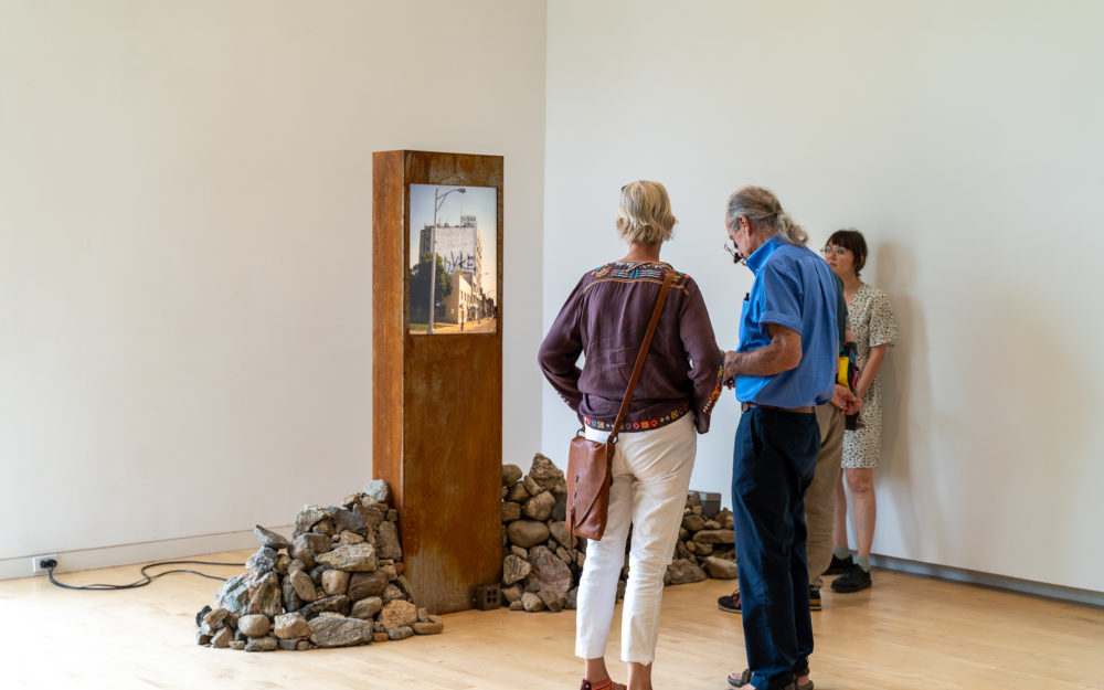 People in a museum gallery looking at an artwork comprised of a lightbox with a photograph surrounded by a stone wall