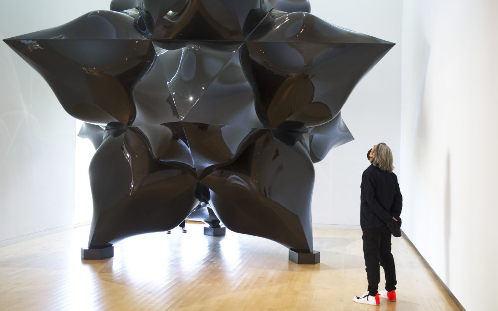 A man wearing all black with long gray hair views a large-scale black star sculpture by Frank Stella.