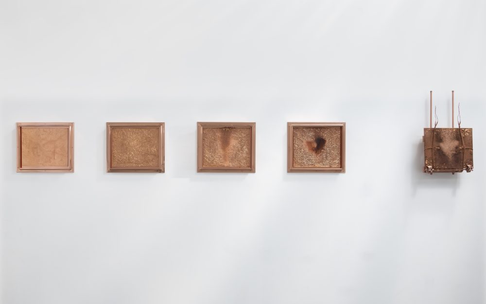 Five etched rectangular copper panels hung on a white wall. The rightmost panel has vertical sculptural elements.