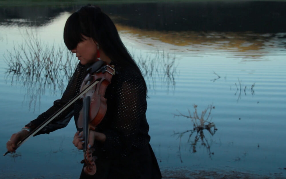 Artist Laura Ortman playing the violin in front of a body of water at dusk