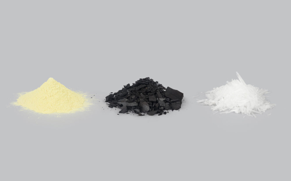 A yellow pile of sulfur, a black pile of charcoal, and a white pile of saltpeter against a gray background