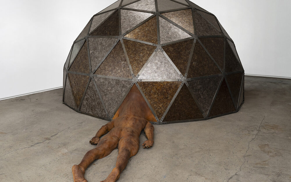 A geodesic dome with a body laying face down on the ground the head obscured by the structure and the bottoms of the feet facing the viewer.