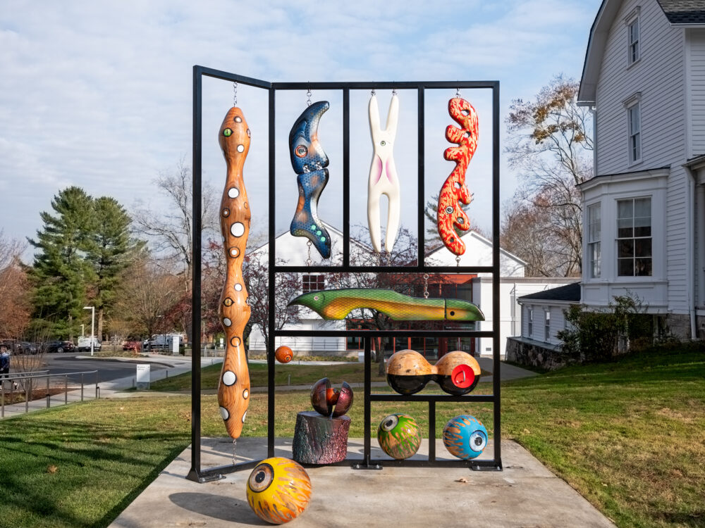 A large-scale sculpture made from steel and salvaged tree matter containing hand-carved, brightly painted objects resembling fishing lures.