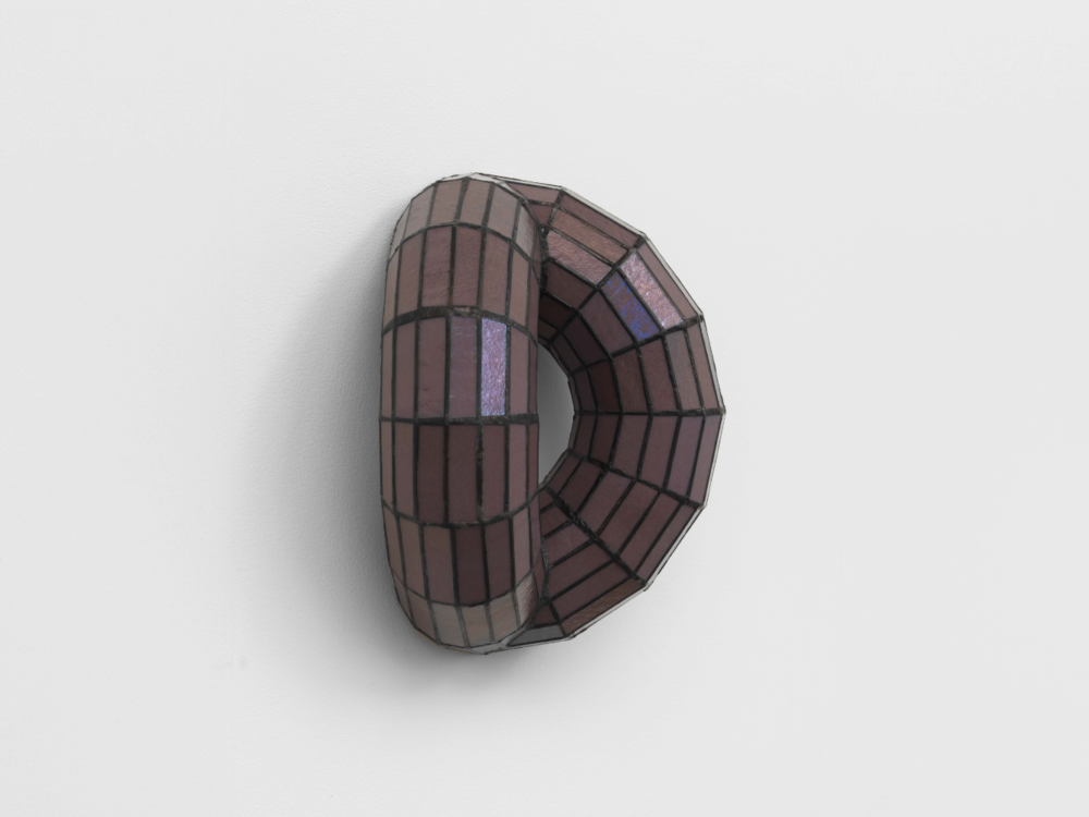 A three dimensional oval stained glass sculpture that's shaped like a ring and bent slightly inward installed on a wall.