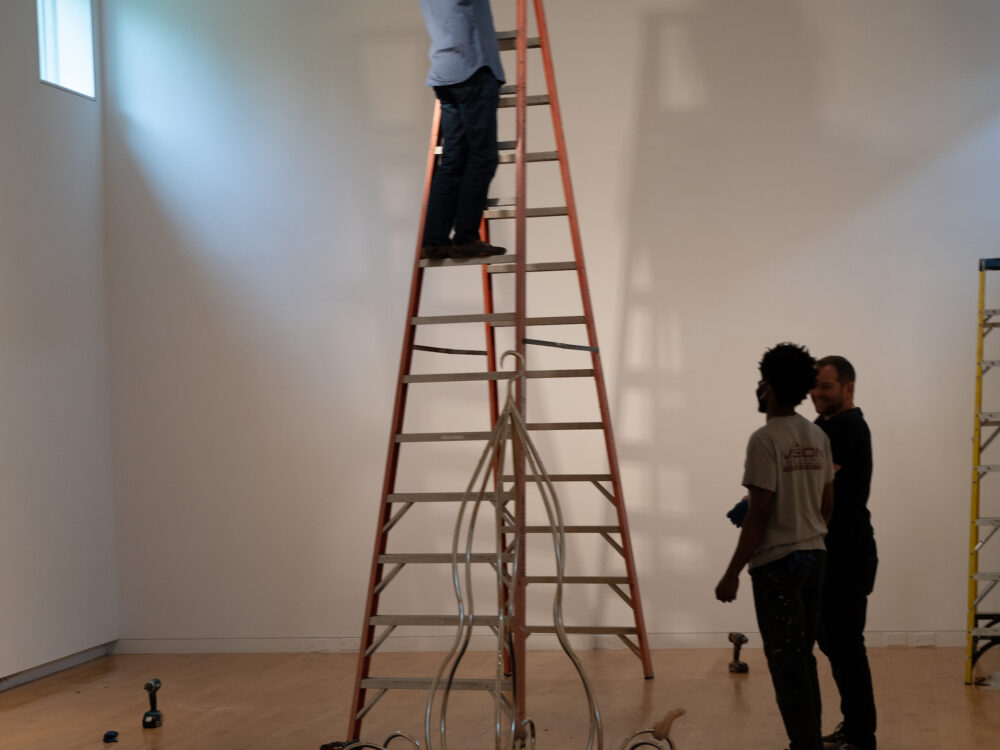 Person on a ladder with chandelier like artwork on a blue blanket with two art handlers nearby.