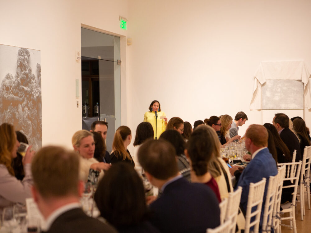 A long table in a gallery with people seated enjoying dinner with a woman wearing yellow giving a speech.