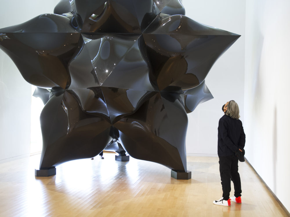 A man wearing all black with long gray hair views a large-scale black star sculpture by Frank Stella.