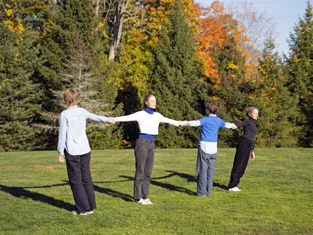Photo of four dancers holding hands in a line with arms outstretched in a field.