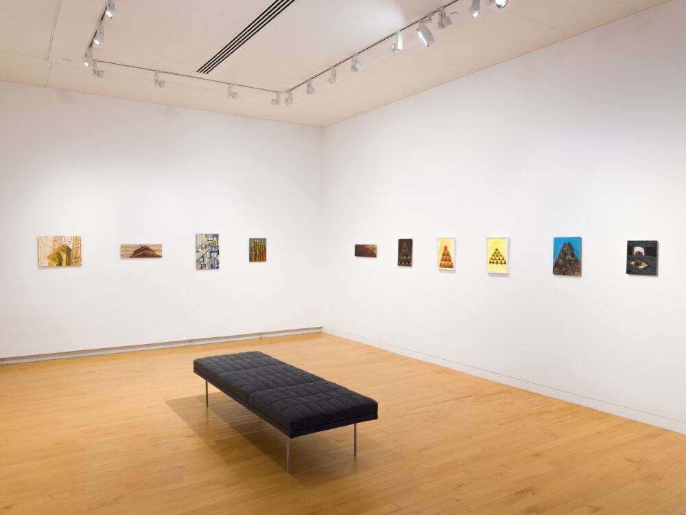 The corner of a room with a bench in the center and a row of small paintings