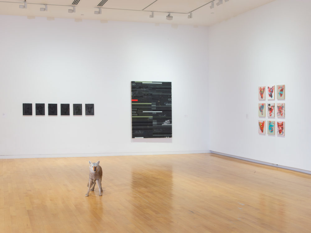 Three abstract paintings on the wall and a model coyote in the center of the gallery.