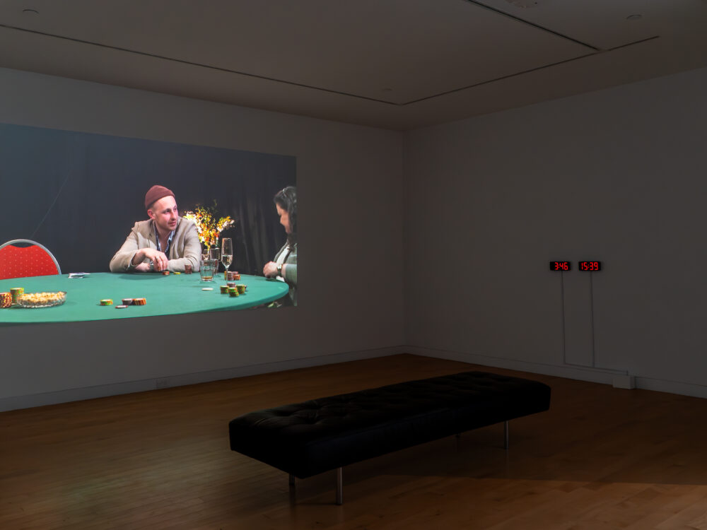 A museum gallery with a video installation projected on a wall and a red timer and clock side-by-side on another wall on the right side