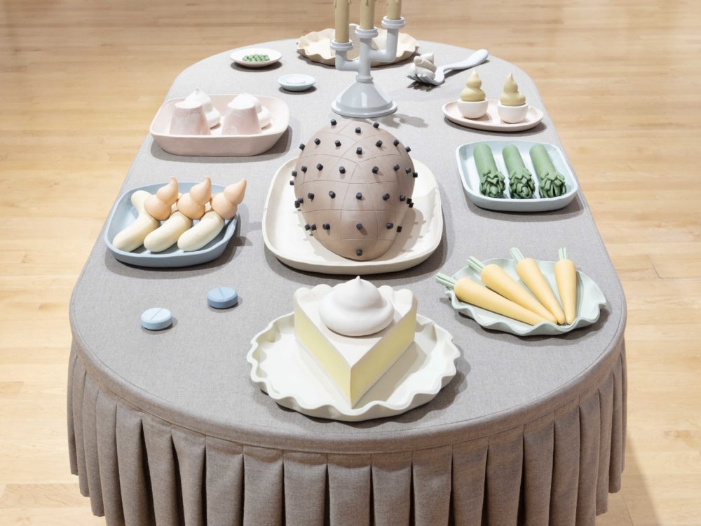 Side view of a oblong table with a gray pleated tablecloth, with clay sculptures of food items such as a ham, slice of pie, deviled eggs, and asparagus.