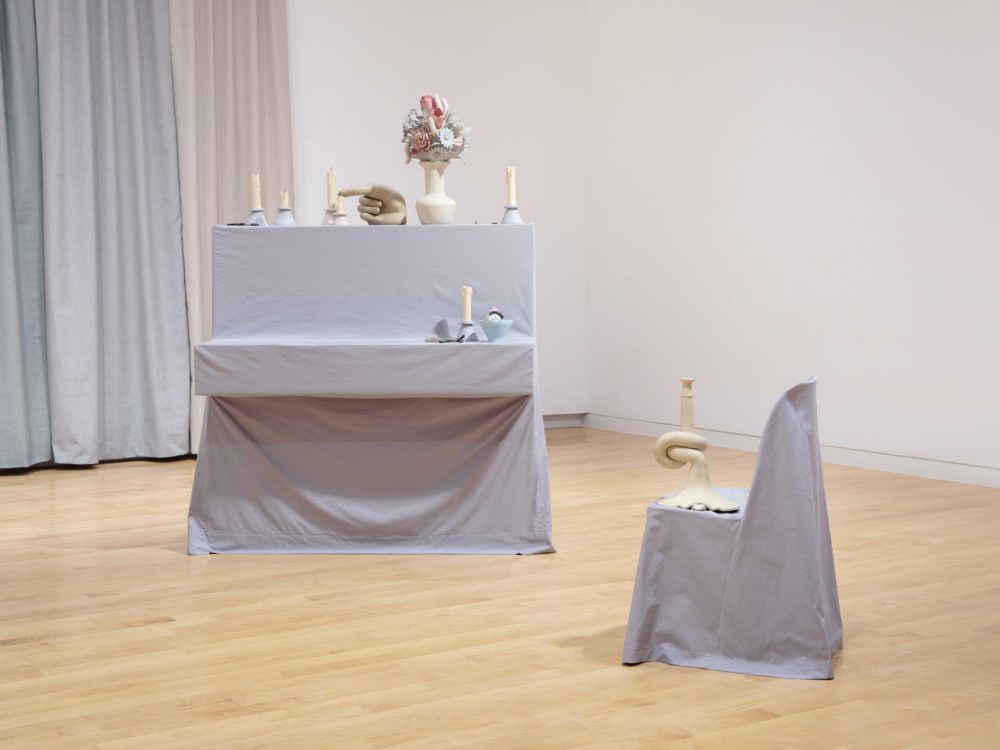 Piano with gray slipcover covered with clay sculptures of extinguished candles and other objects and a side view of a chair with a gray slipcover.