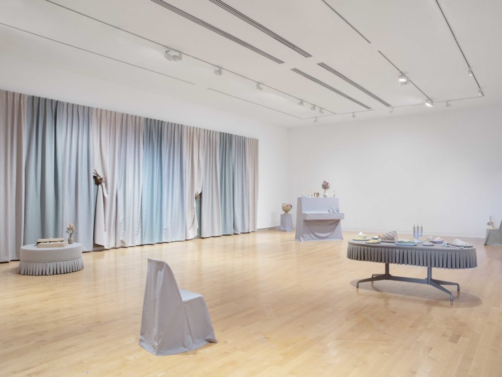 Overall view with several gray slipcovered furnishings with small surrealist clay objects and a large-scale multi-colored curtain installation to the left.