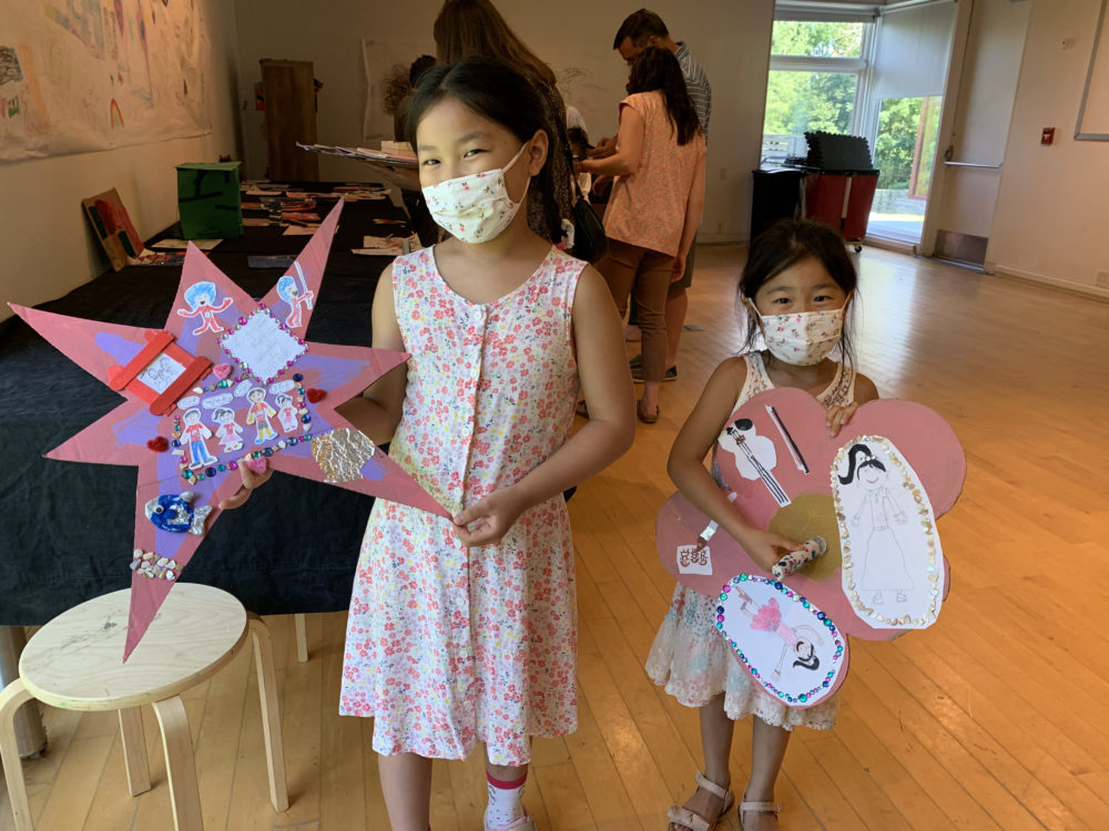 Two young girls holding their artwork. One holds a pink star collage and the other holds a pink flower collage.