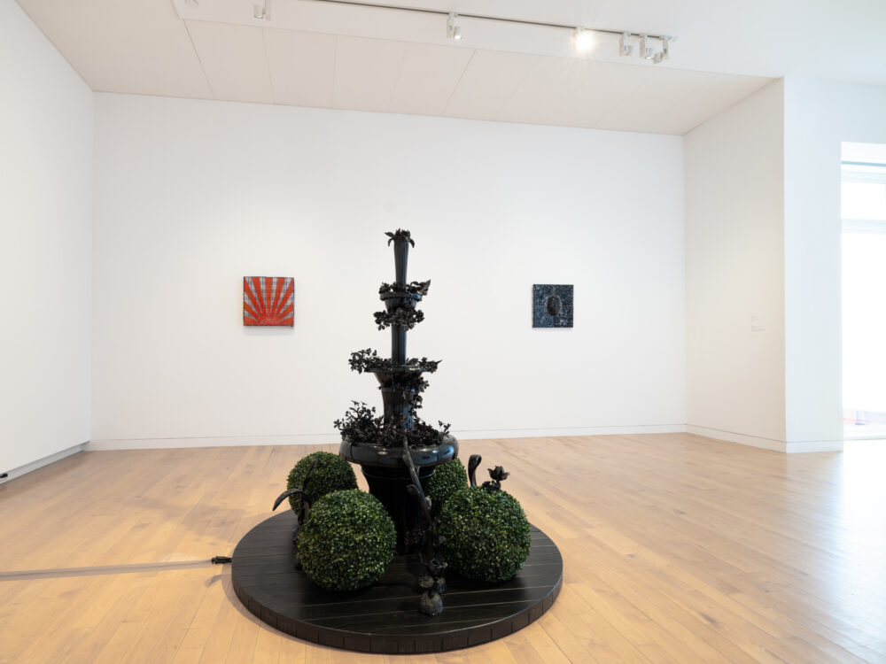 A black glass fountain in the center of the room with two small wall mounted works in the background.
