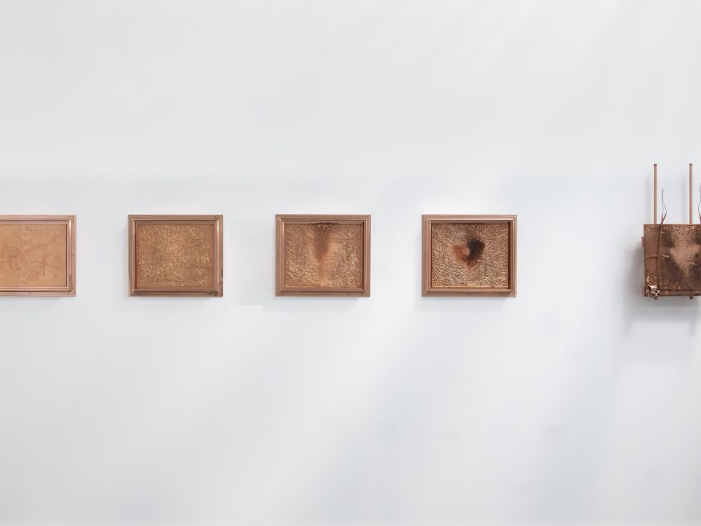 Five etched rectangular copper panels hung on a white wall. The rightmost panel has vertical sculptural elements.