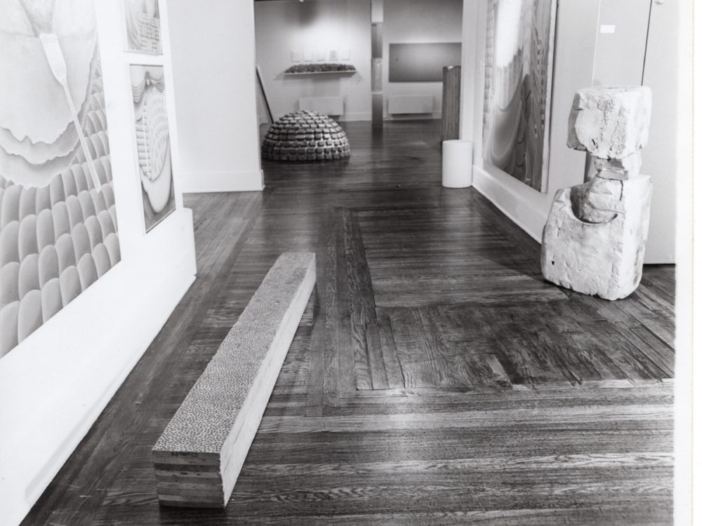 Black and white photo of a gallery with paintings on a wall to the left a low long rectangular sculpture and an abstract upright sculpture to the right.
