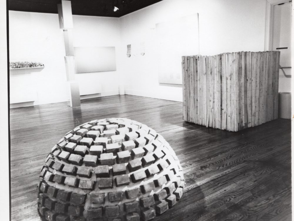 Black and white photo with brick dome sculpture in foreground, wooden cube-like structure in background and paintings on the walls.