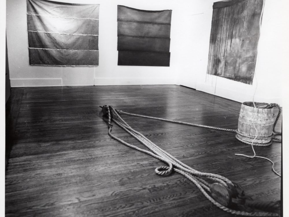 Black and white photo with a sculpture comprised of a bucket and rope on the floor with unstretched abstract works on the wall.