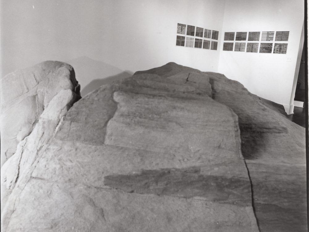 Black and white photo with grids of small works on the walls in the background and a large abstract floor sculpture that looks like mountainous terrain.