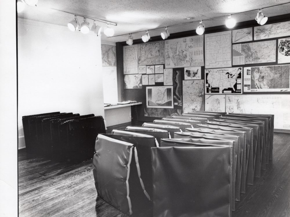 Black and white photo with an accordion-folded large floor sculpture in the foreground and many works on paper hung on a wall in the background.