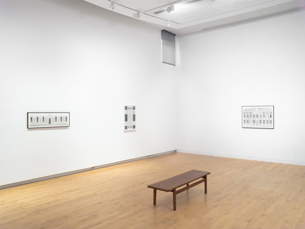 Installation view of a corner of a gallery with three framed drawings on the walls and a wooden bench in the center of the room.