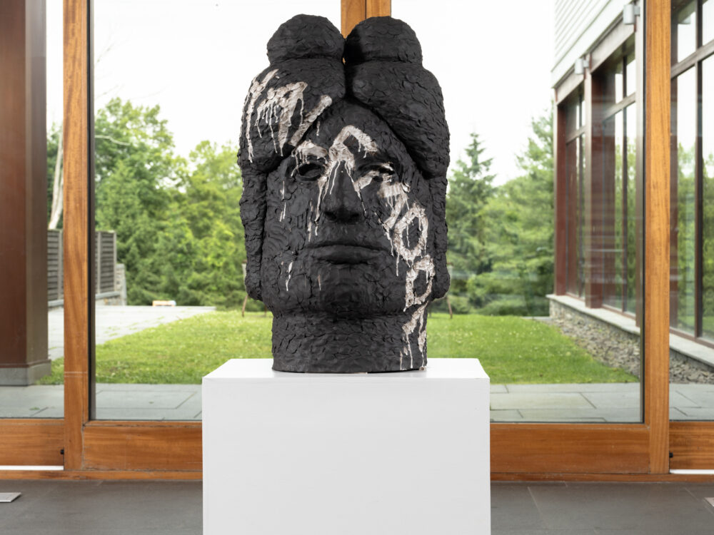 Large ceramic sculpture of a head on a white pedestal in front of glass windows.