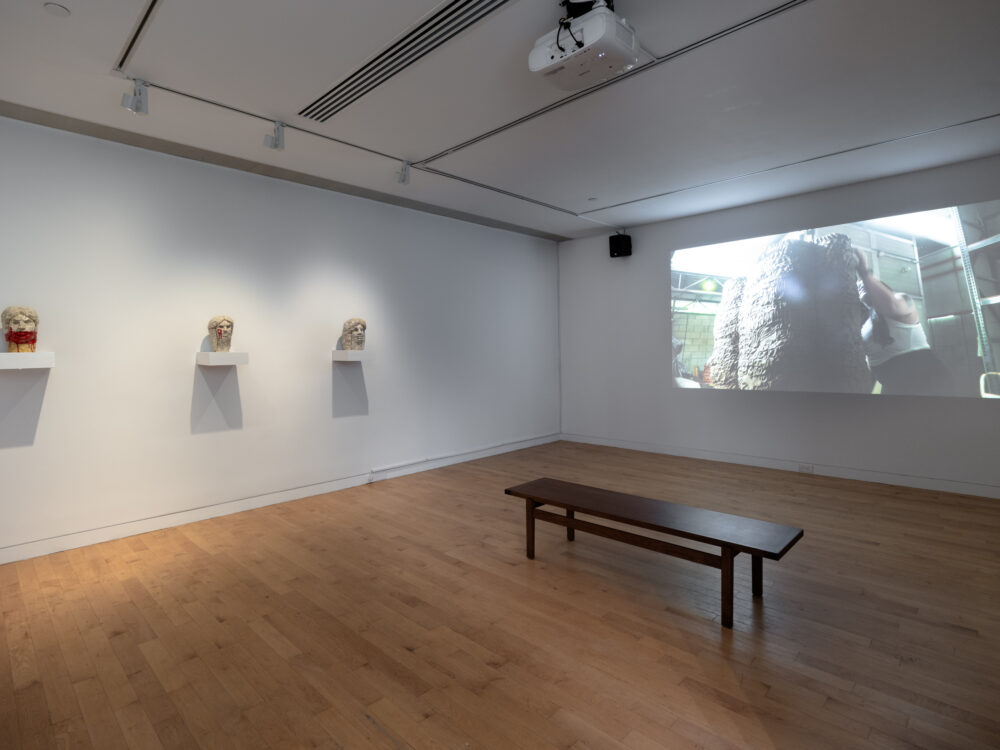 Three small sculptures of heads on shelves to the left and a video projection to the right.
