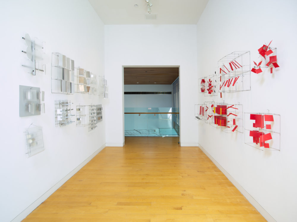 Gallery space with a doorway at center. To the right are small red material and wire sculptures hung on the wall. To the left are small reflective sculptures.