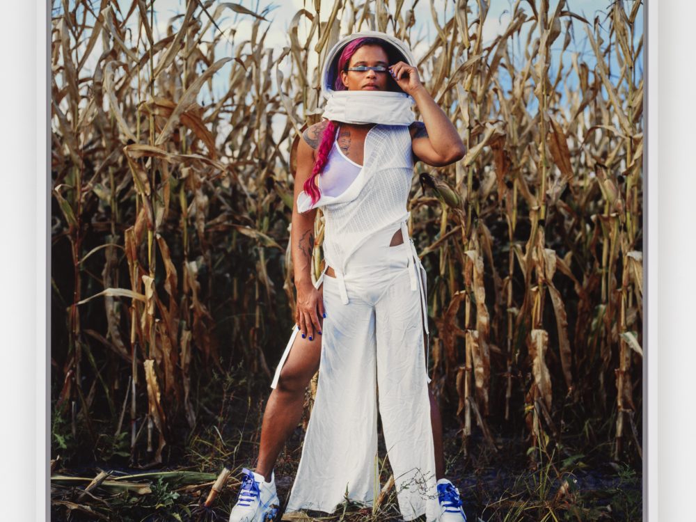 Photograph of a person wearing sneaker anda helmet-like headpiece, with a long fuchsia braid standing in a cornfield.