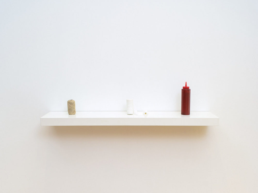 A roll of twine, roll of white bandages, and a red bottle of ketchup sit on a white shelf in gallery